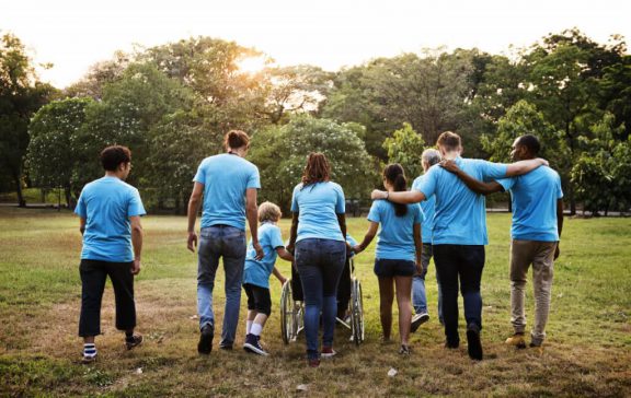 A group of people of varying ages, races and abilities are gathered in a meadow with their arms around each other.