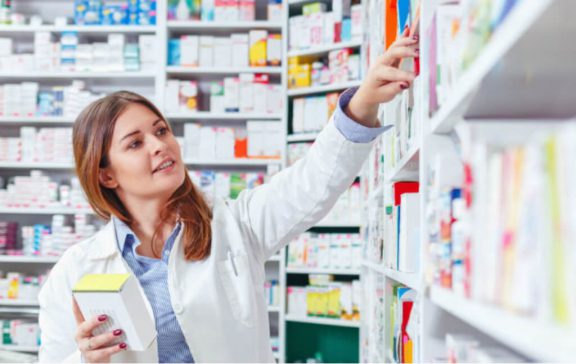 A female pharmacists in a white coat reaches for medication on a pharmacy shelf.