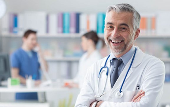 A male doctor wearing a white coat and stethoscope smiles. Two other people are blurred in the background.